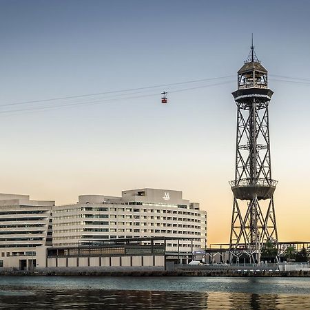 Eurostars Grand Marina Hotel GL Barcelona Exterior foto The photo shows a modern building alongside a waterfront, with a distinctive tower nearby. The tower appears to be part of a cable car system, featuring a conical roof and a series of cables extending from it. The scene is set against a colorful sky,
