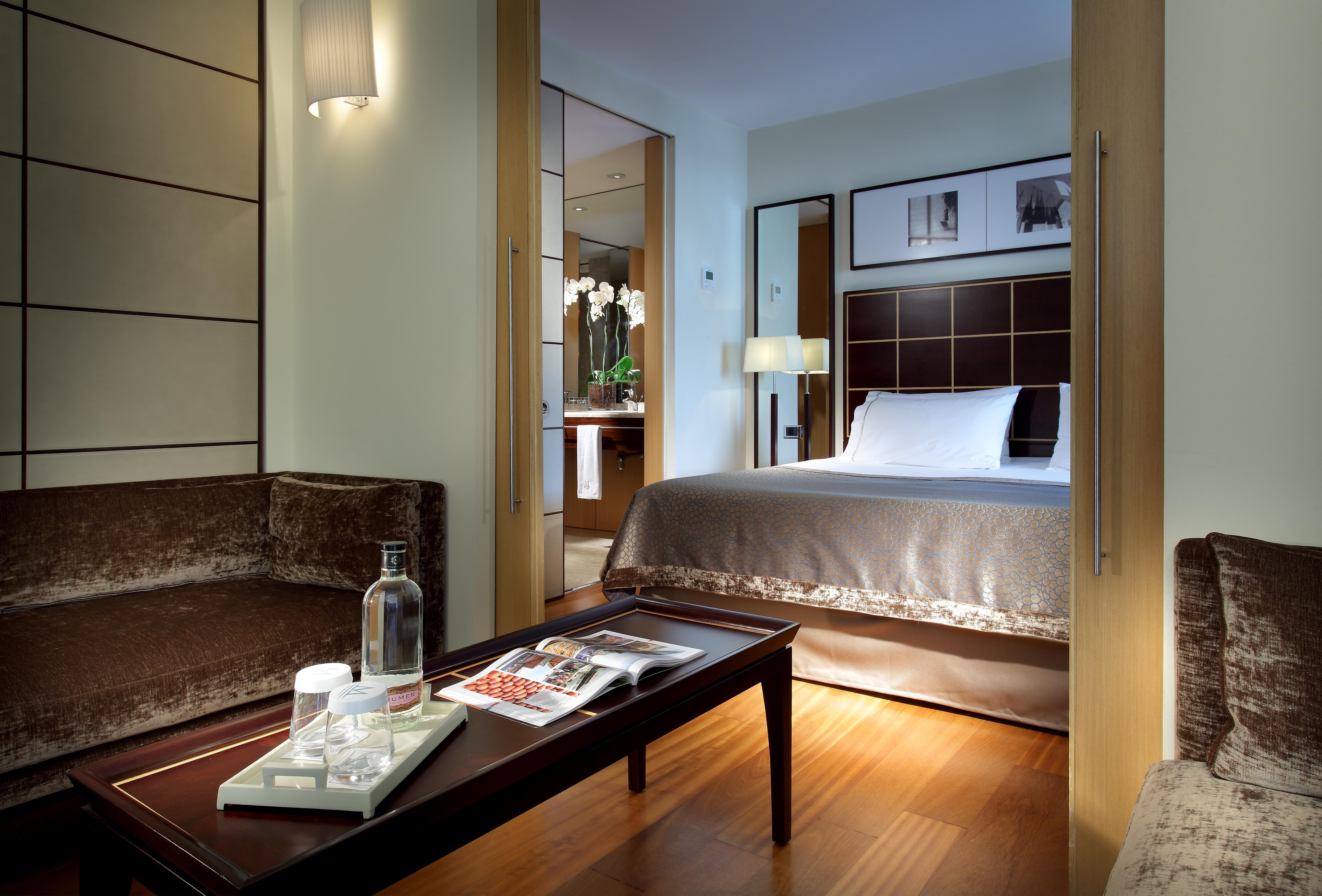 Eurostars Grand Marina Hotel GL Barcelona Exterior foto The image shows a well-decorated hotel room featuring a comfortable bed with a plush headboard, complemented by a subtle color palette. There is a cozy seating area with a brown couch and a coffee table. On the table, there are items like a water bot