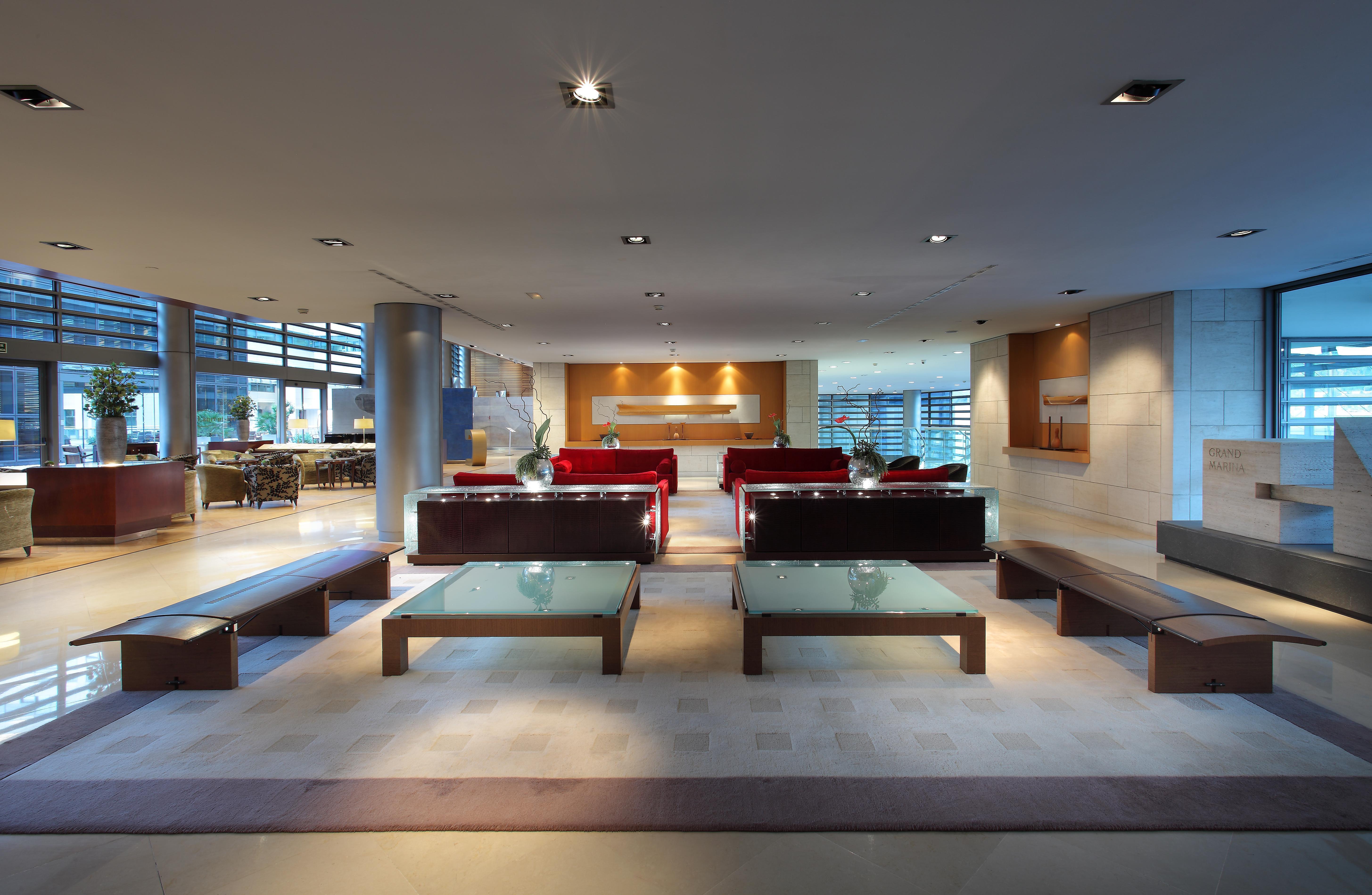 Eurostars Grand Marina Hotel GL Barcelona Exterior foto The photo shows a modern and spacious interior, likely of a lobby or waiting area. It features several red sofas arranged in a conversational layout, complemented by glass-top coffee tables. The lighting is bright and there are large windows, allowin