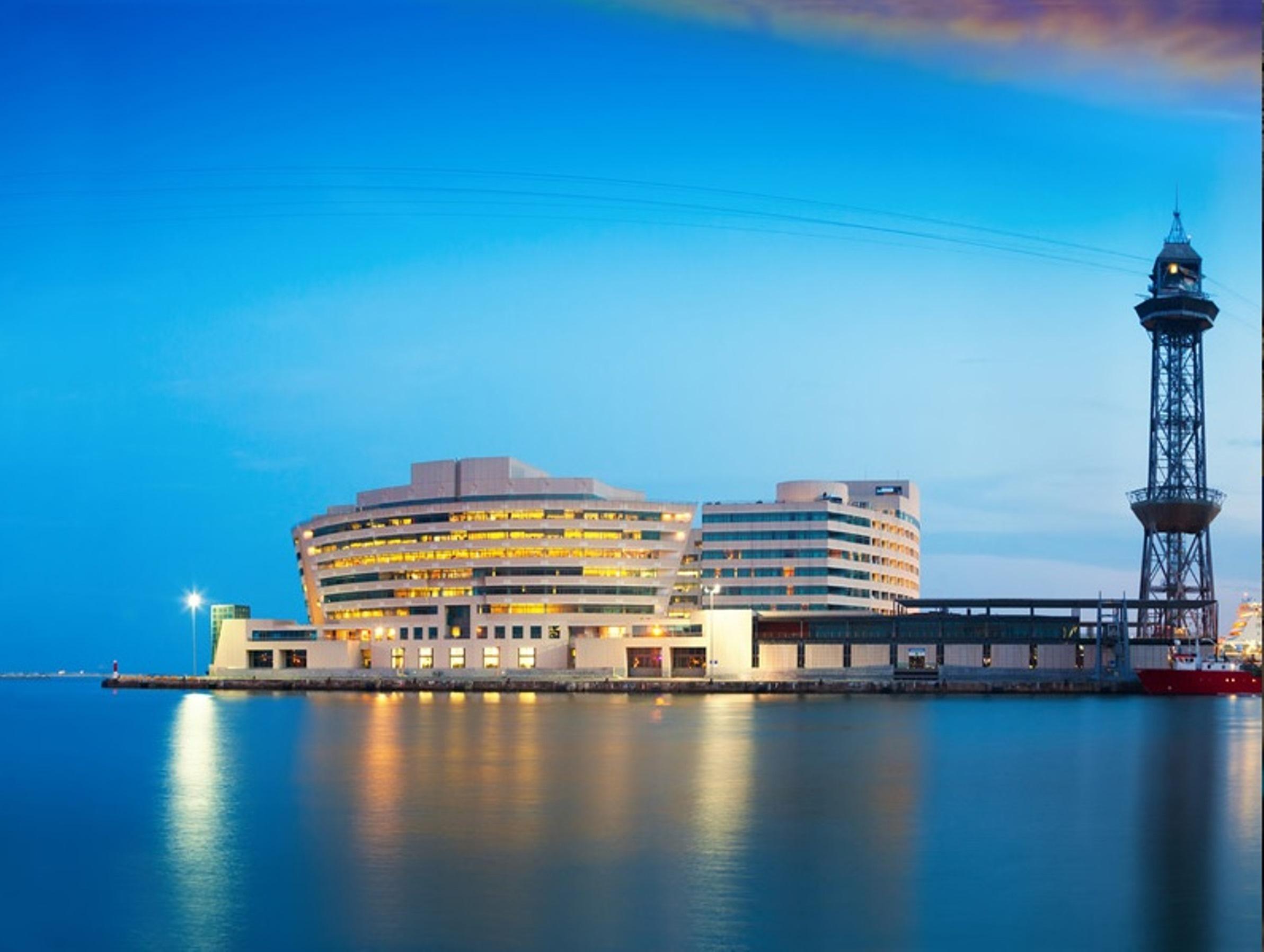 Eurostars Grand Marina Hotel GL Barcelona Exterior foto The photo shows a modern building situated near the water, likely a harbor or port area. The structure features a sleek, curved design with multiple levels and large windows, reflecting an urban architectural style. Next to the building, there is a t