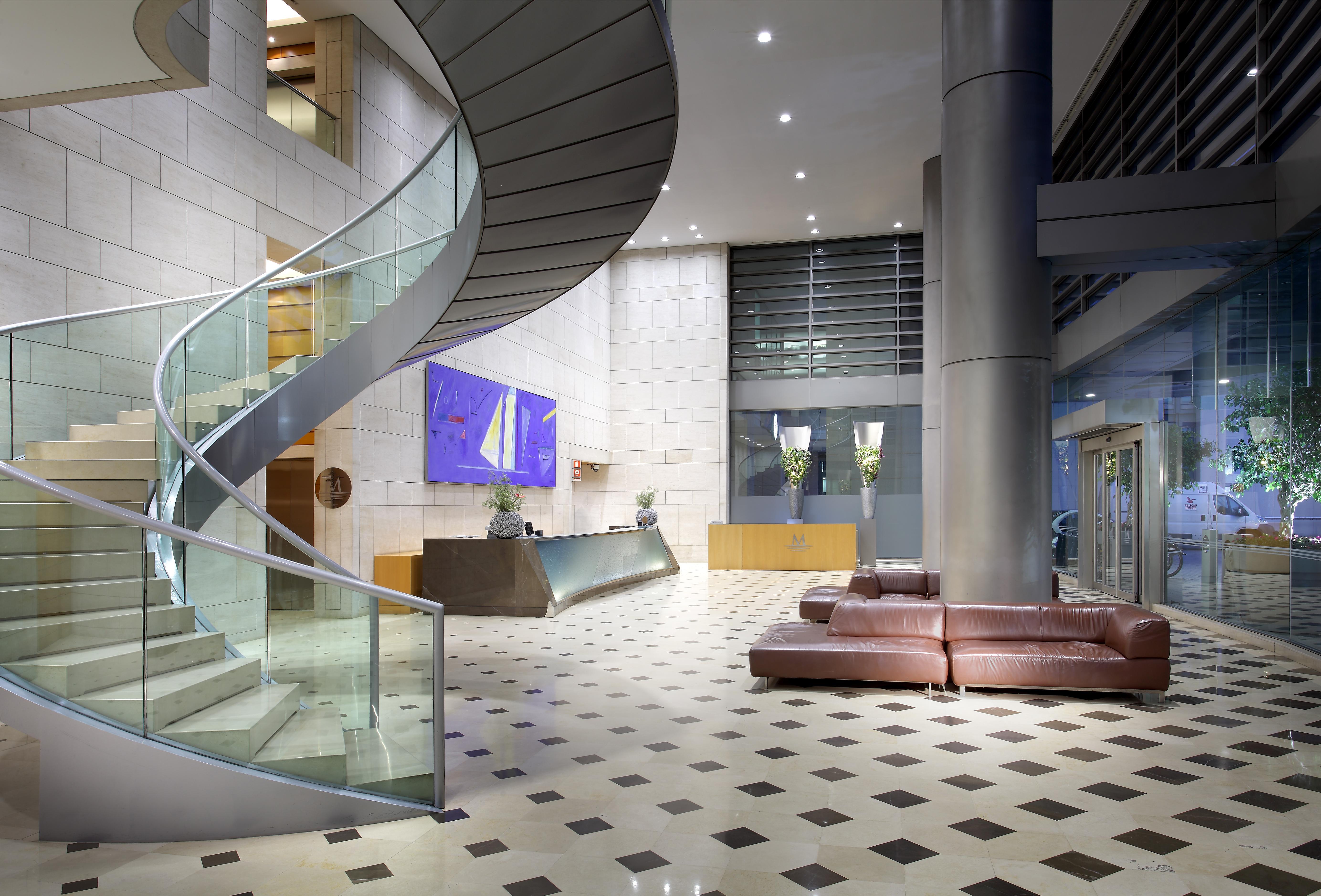 Eurostars Grand Marina Hotel GL Barcelona Exterior foto The photo shows a modern hotel or office lobby featuring a sleek, spiral staircase made of glass and metal. The staircase is accompanied by a large, open space with a tiled floor that has a checkered pattern, combining light and dark tiles. 

To the 