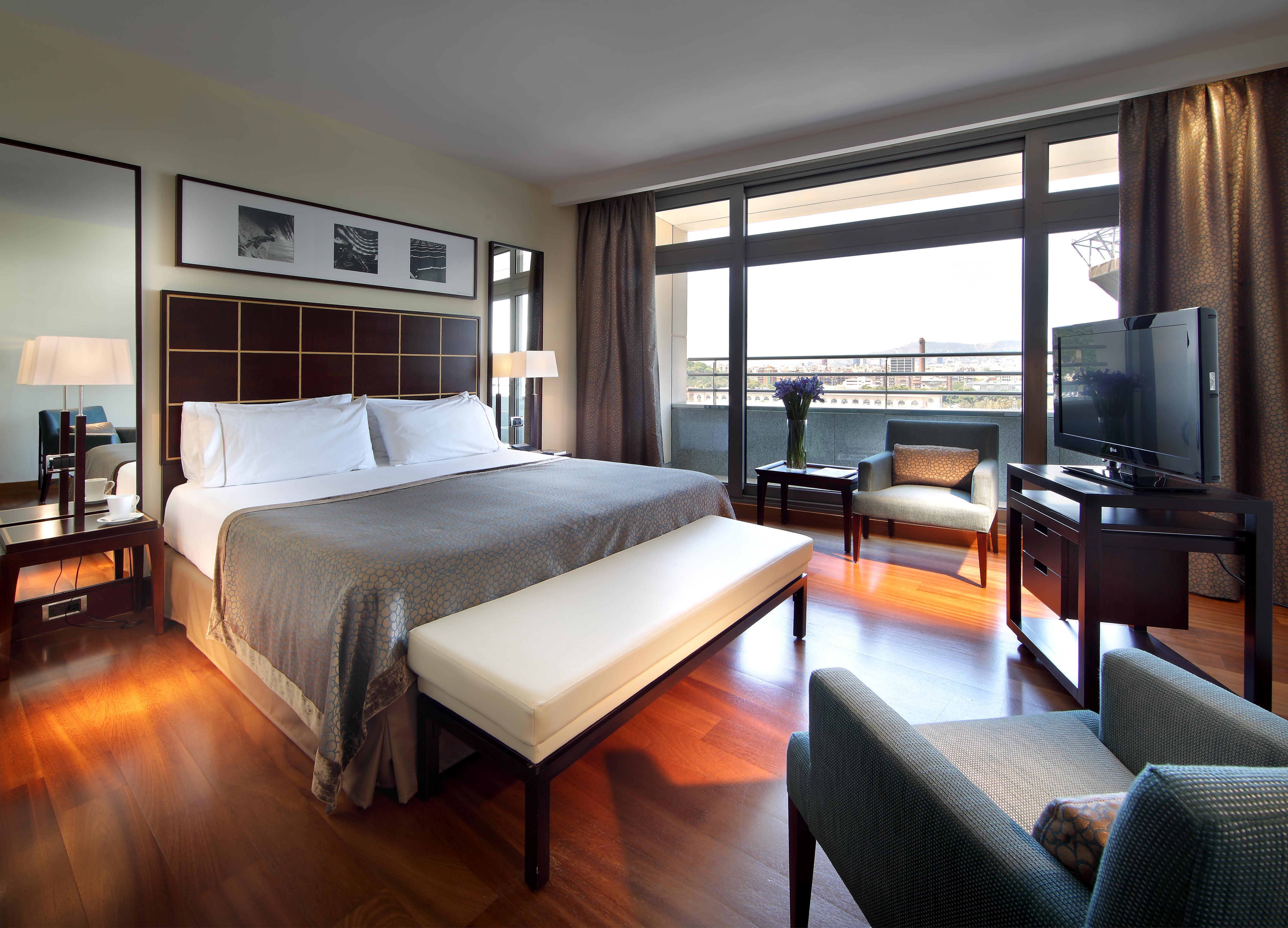 Eurostars Grand Marina Hotel GL Barcelona Exterior foto The photo shows a cozy hotel room featuring a large bed with white linens and a gray bedspread. There is a light-colored bench at the foot of the bed. A small seating area includes a comfortable chair and a lamp, along with a wooden side table. The r