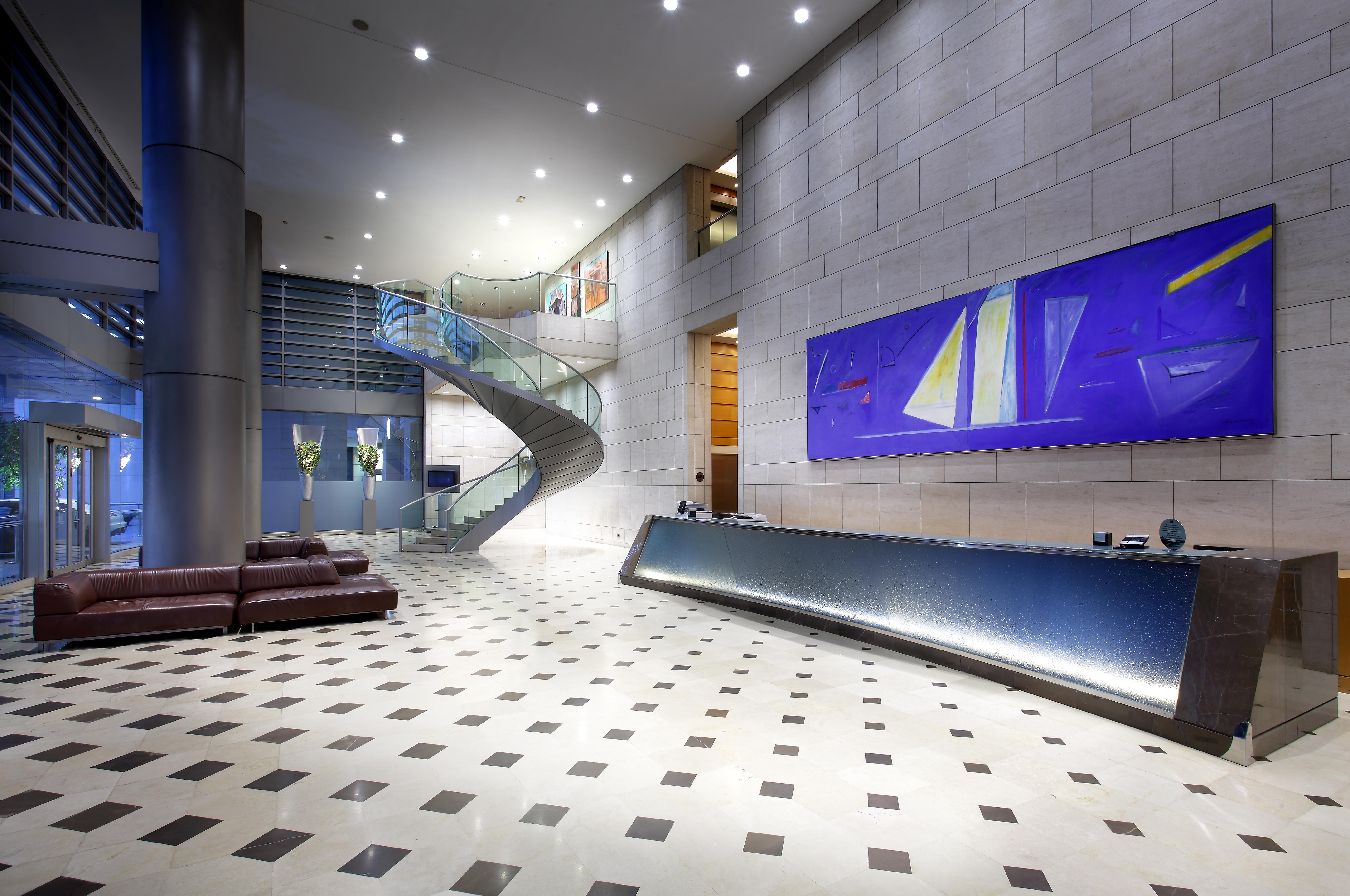 Eurostars Grand Marina Hotel GL Barcelona Exterior foto The photo shows a modern lobby or reception area. There is a sleek reception desk along one wall, featuring a polished surface. To the left, a spacious staircase with a glass guardrail leading to an upper level can be seen, framed by walls decorated 