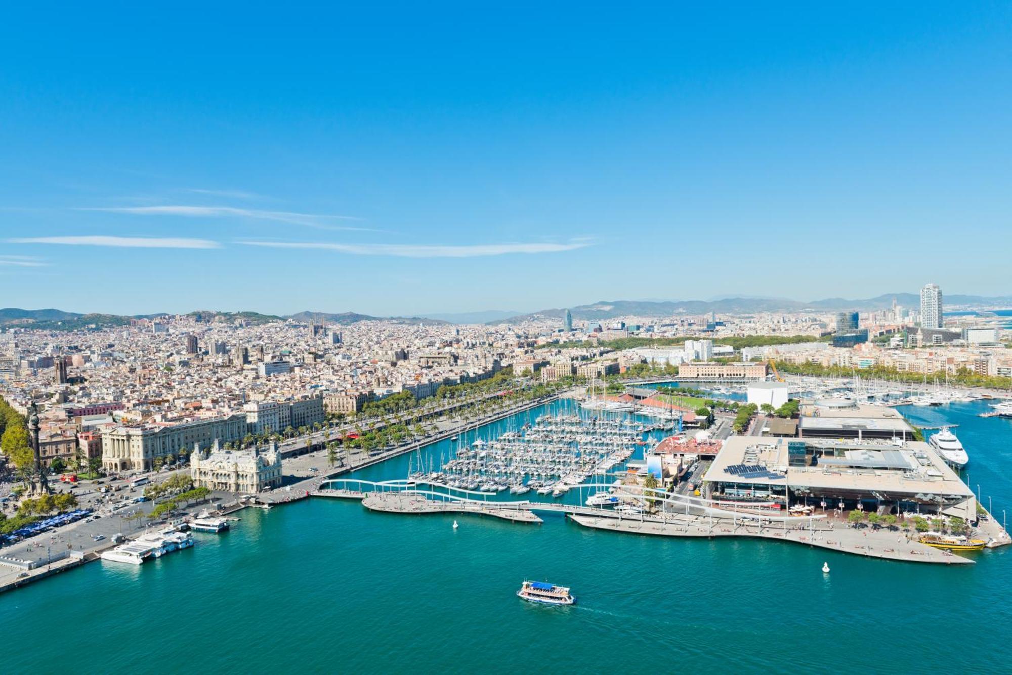 Eurostars Grand Marina Hotel GL Barcelona Exterior foto The photo depicts a coastal city view, likely of a vibrant harbor or marina. In the foreground, there is a clear blue body of water with a few small boats. The harbor features numerous yachts and boats docked in the marina. 

Surrounding the water ar