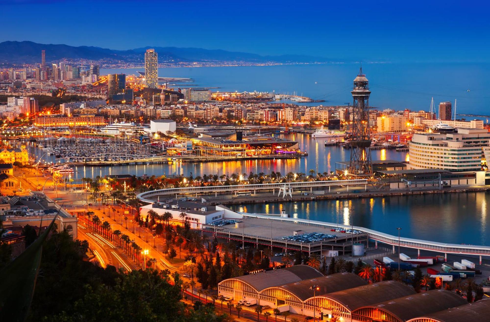 Eurostars Grand Marina Hotel GL Barcelona Exterior foto The photo depicts a scenic view of a coastal city at dusk. In the foreground, there are illuminated buildings and a marina filled with boats. The waterfront features a curved walkway and a dock area. In the background, you can see a skyline with tall