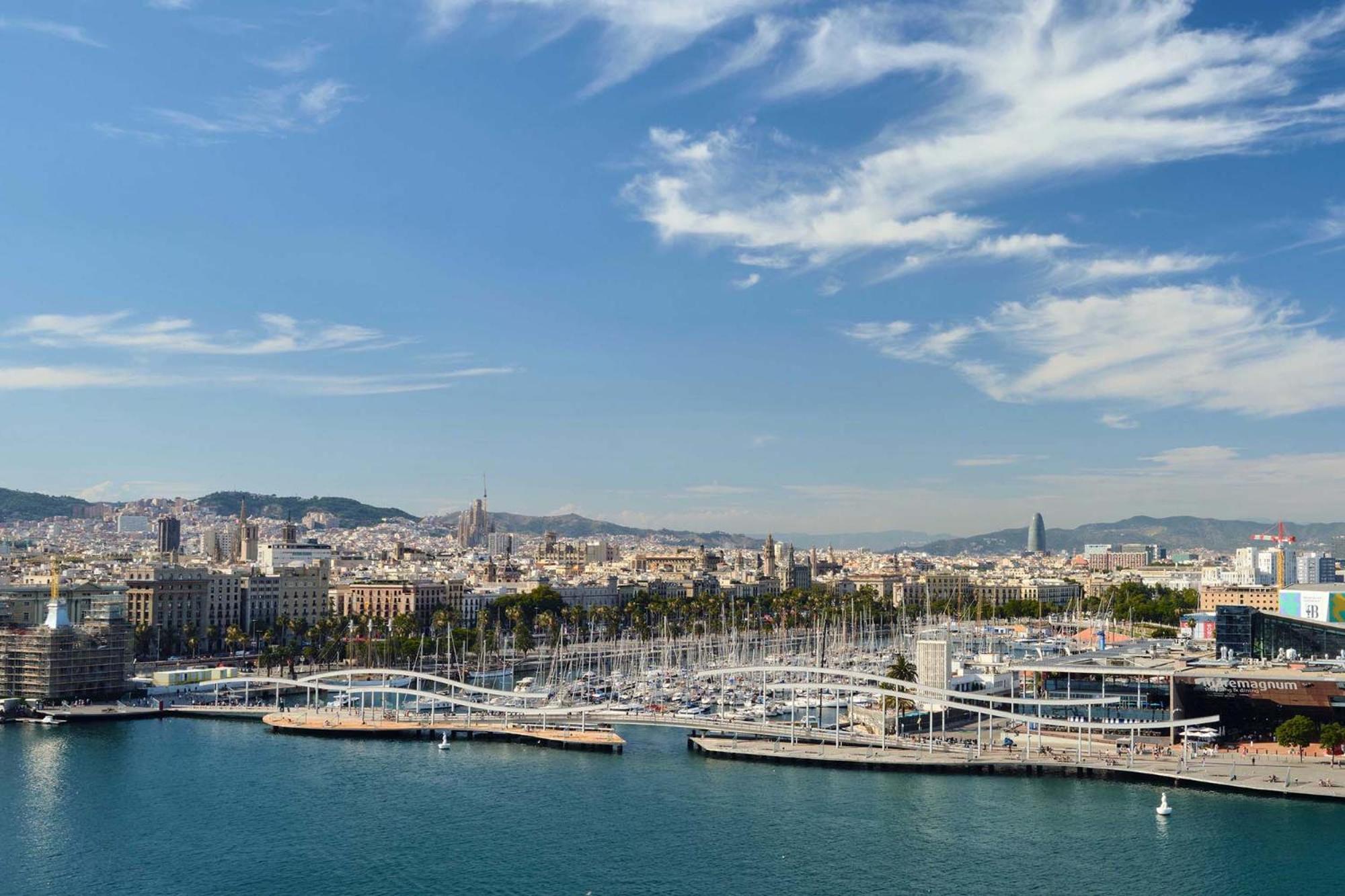 Eurostars Grand Marina Hotel GL Barcelona Exterior foto The photo shows a scenic view of a coastal city, likely Barcelona, with a harbor area prominent in the foreground. There are several boats moored along the docks, and a distinctive bridge or walkway structure can be seen connecting different parts of