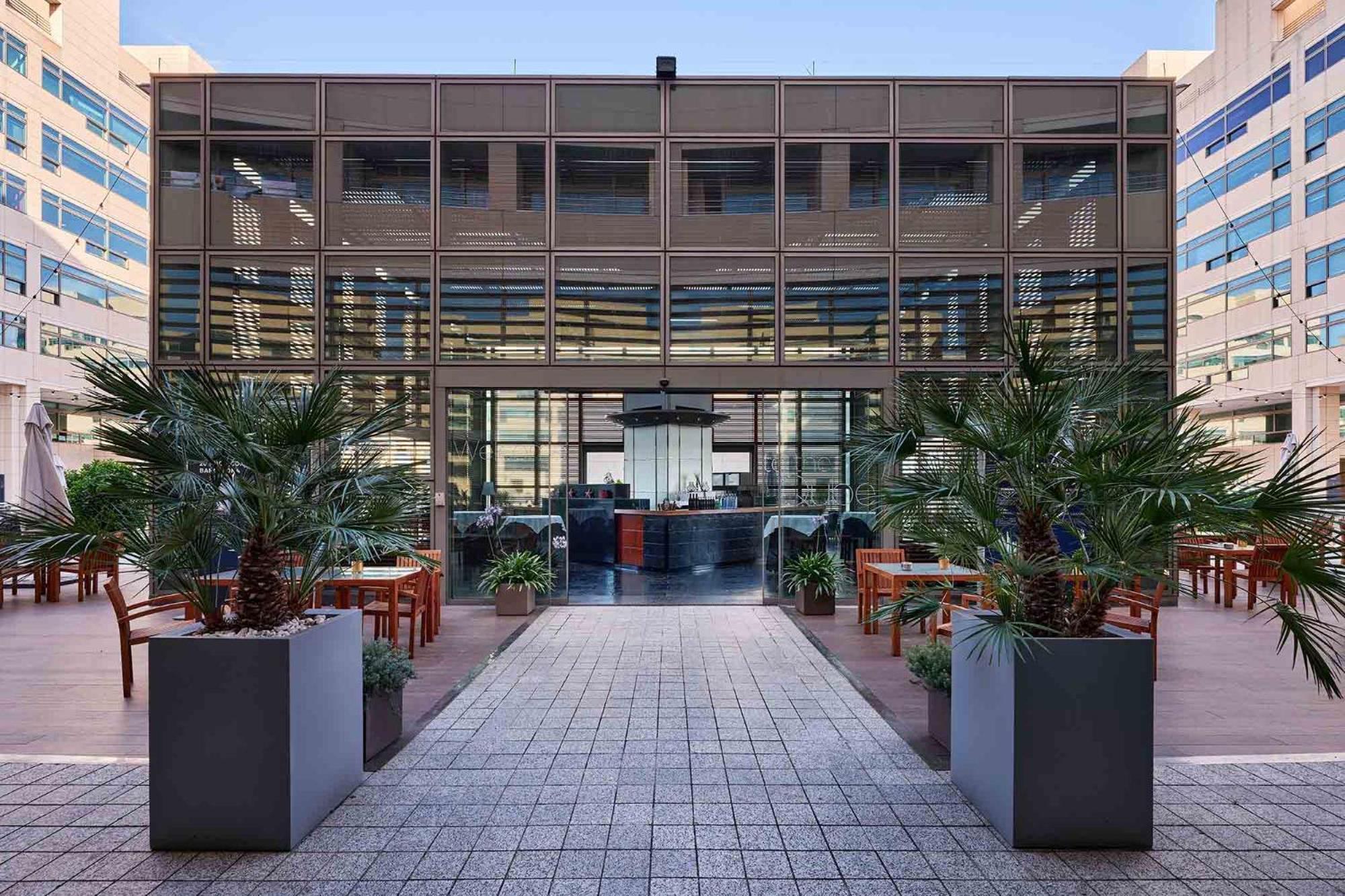 Eurostars Grand Marina Hotel GL Barcelona Exterior foto The photo depicts a modern building with a glass facade, featuring a central entrance framed by large planters with tropical plants. The entrance leads to an interior space that appears to be a restaurant or café, with a stylish counter and seating v