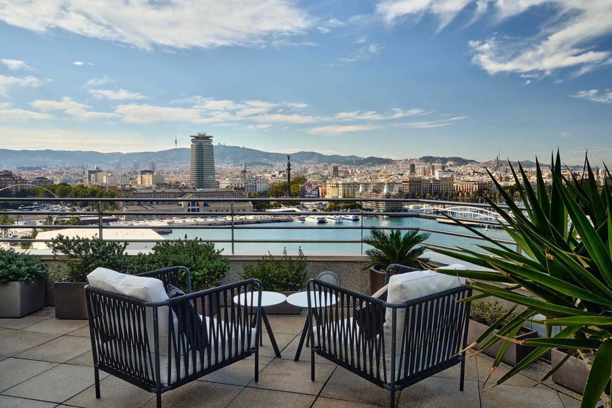 Eurostars Grand Marina Hotel GL Barcelona Exterior foto The photo shows a scenic outdoor terrace area featuring two stylish black chairs with white cushions. In the background, there’s a beautiful view of a cityscape with a harbor and mountains in the distance. The sky is partly cloudy, and there are some