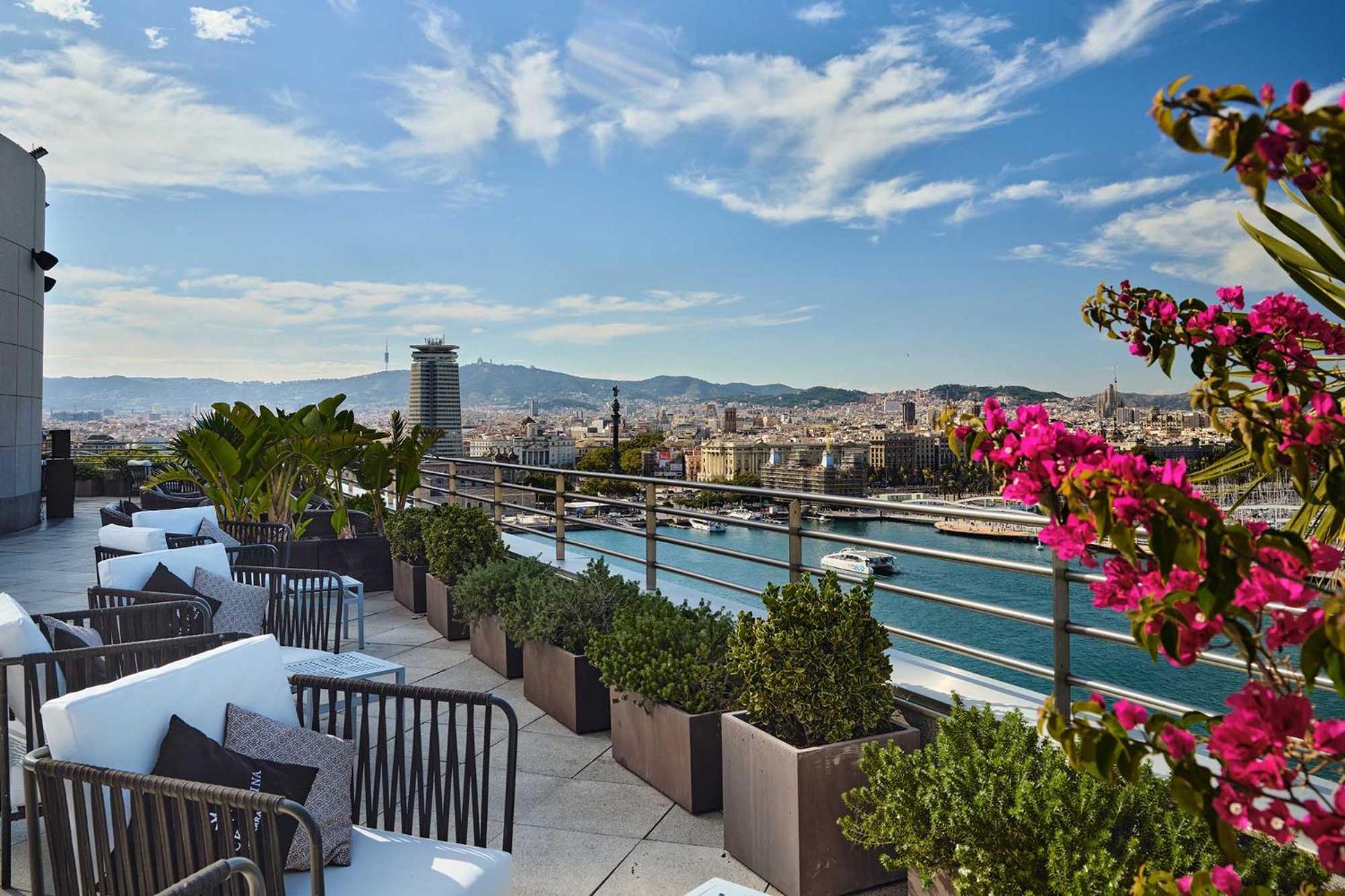 Eurostars Grand Marina Hotel GL Barcelona Exterior foto The photo depicts a rooftop terrace with a stunning view of a city, likely Barcelona. There are comfortable seating arrangements, including dark outdoor chairs and cushions, surrounded by decorative plants in large pots. Colorful flowers, possibly bo