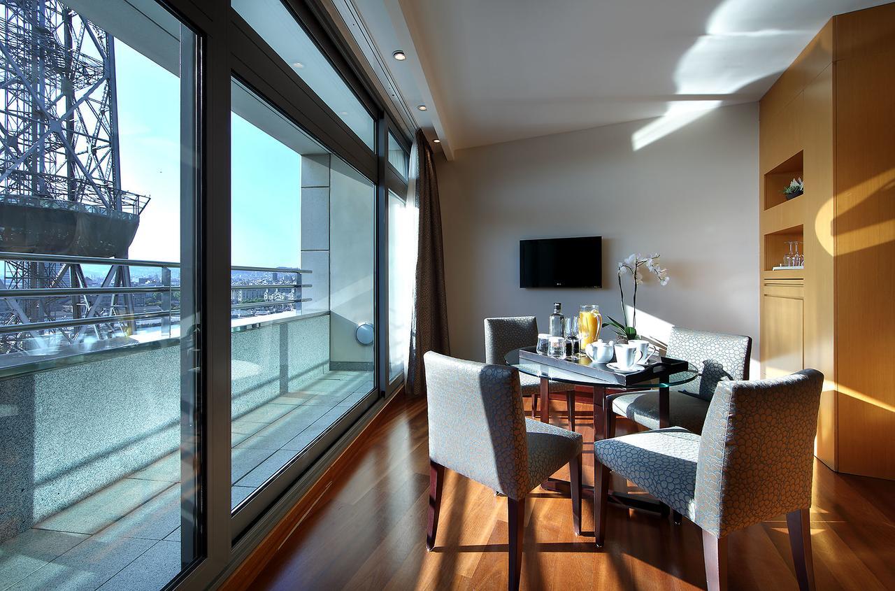 Eurostars Grand Marina Hotel GL Barcelona Exterior foto The photo shows a modern and bright room that features large windows with panoramic views. Inside, there is a circular dining table surrounded by upholstered chairs. On the table, there are items that suggest a meal or refreshments, such as drinks an