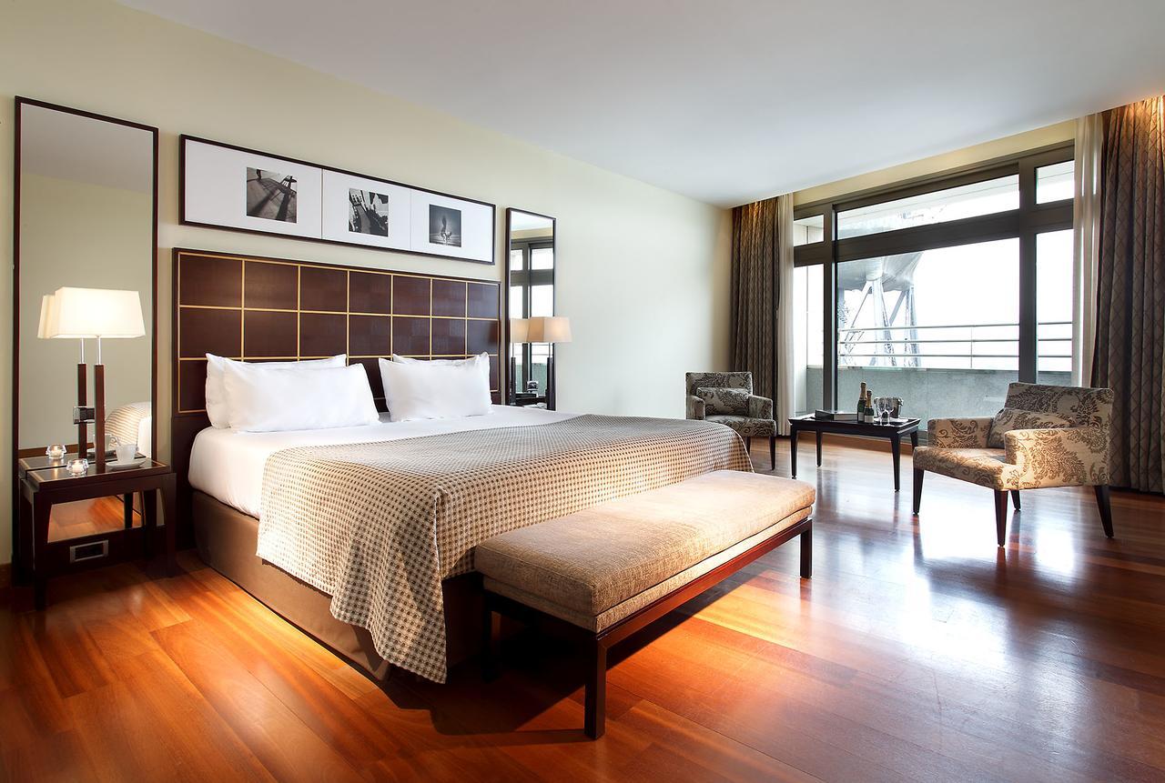 Eurostars Grand Marina Hotel GL Barcelona Exterior foto The photo shows a modern hotel room featuring a large bed with a decorative headboard. There are two bedside lamps on either side of the bed, and behind the bed, there are framed pictures hanging on the wall. A bench sits at the foot of the bed, and 