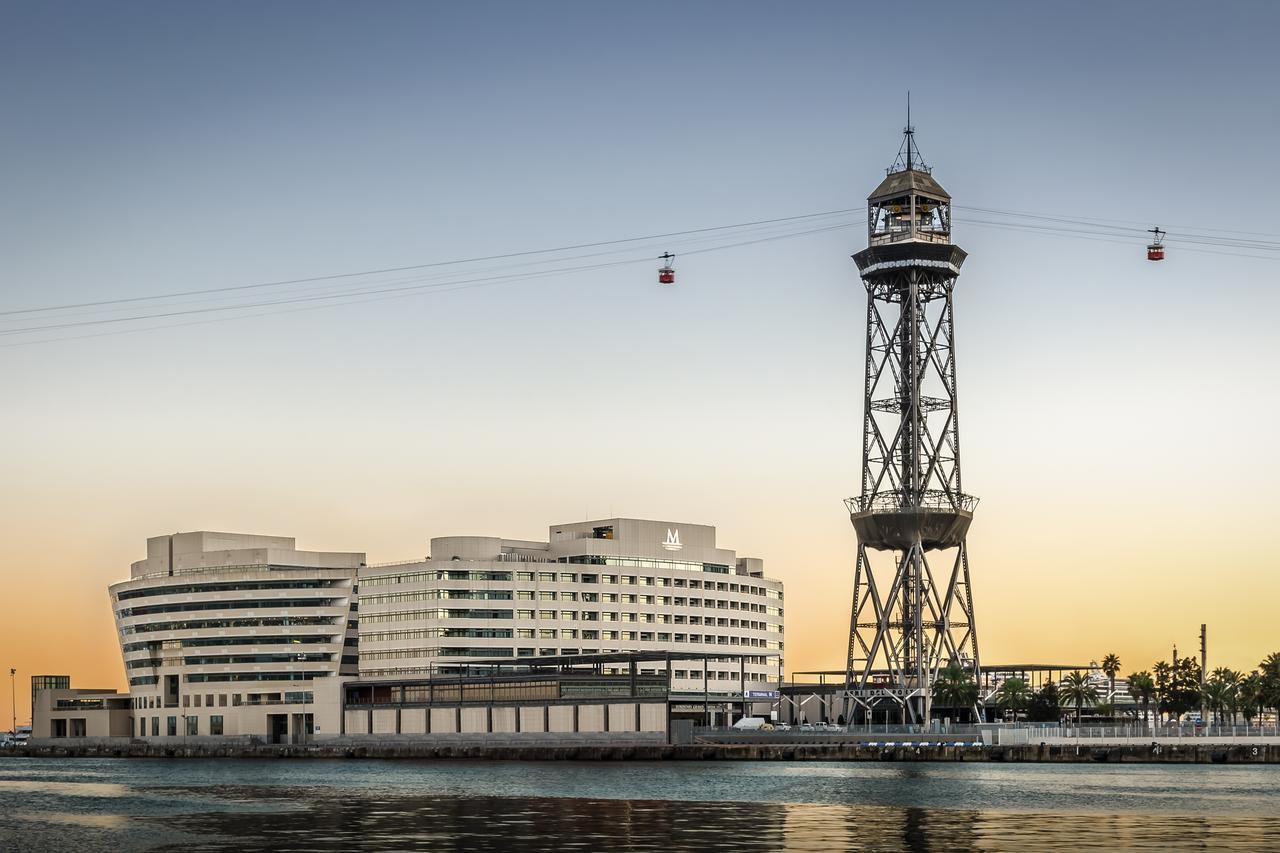 Eurostars Grand Marina Hotel GL Barcelona Exterior foto The photo shows a modern building alongside a waterfront, with a distinctive tower nearby. The tower appears to be part of a cable car system, featuring a conical roof and a series of cables extending from it. The scene is set against a colorful sky,