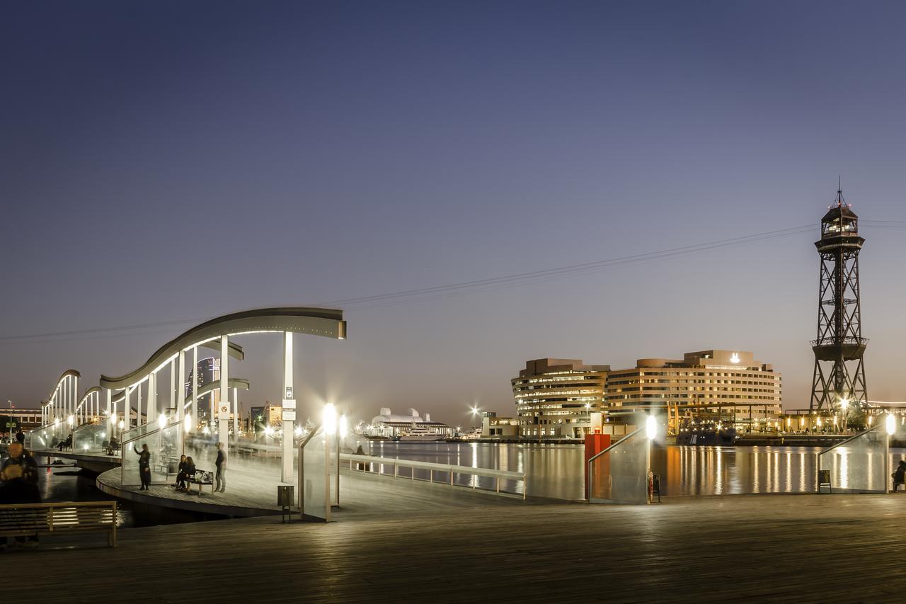 Eurostars Grand Marina Hotel GL Barcelona Exterior foto The photo depicts a scenic waterfront view during twilight. On the left, there is a modern, curved pedestrian walkway that appears to be well-lit, inviting pedestrians. To the right, a tall structure resembling a lighthouse or observation tower stand